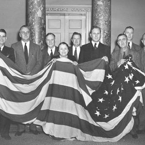 Group of older white men in suits and white women holding up large American flag in room with columns
