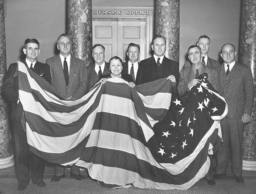 Group of older white men in suits and white women holding up large American flag in room with columns
