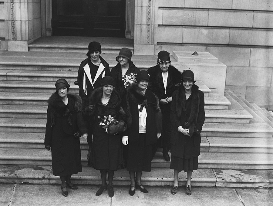 Group of seven white women standing on marble stairs in dresses coats and hats
