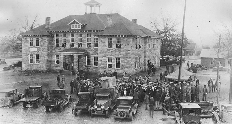 Crowd of people standing with cars and trucks parked outside multistory stone building with cupola