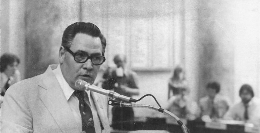 White man in suit with glasses speaking at lectern