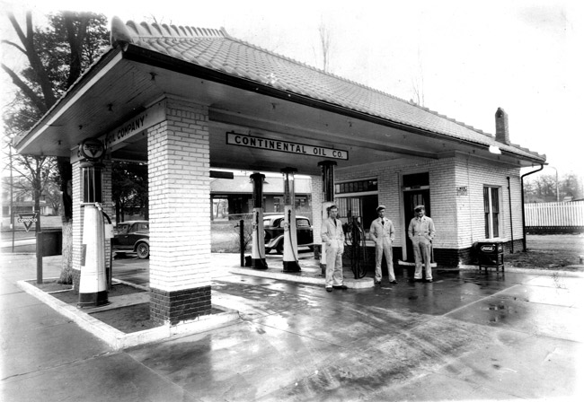 Three white male workers standing near pumps at a brick gas station under sign saying "continental oil co."