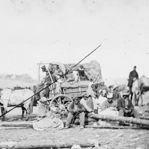 African-Americans in covered wagon and sitting on logs with white soldiers on horseback behind them