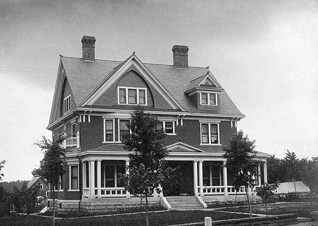 Black and white photo of three-story brick house with covered porch and brick chimneys and small trees in front