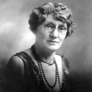 Older white woman with glasses wearing a beaded necklace and fancy dress