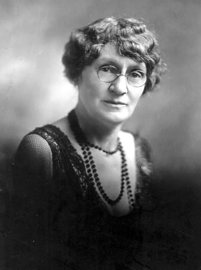 Older white woman with glasses wearing a beaded necklace and fancy dress