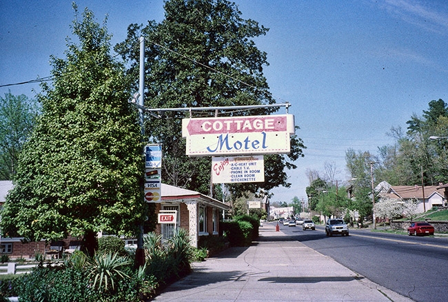 building on street with houses and cars and hanging "cottage motel" sign