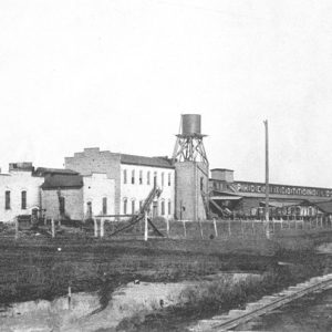 Industrial buildings with smokestack and water tower and rail line in foreground