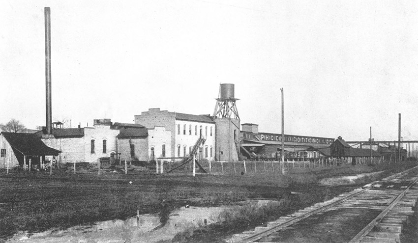 Industrial buildings with smokestack and water tower and rail line in foreground