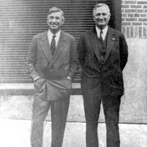 Two men in suit and tie pose for photograph with brick wall behind them