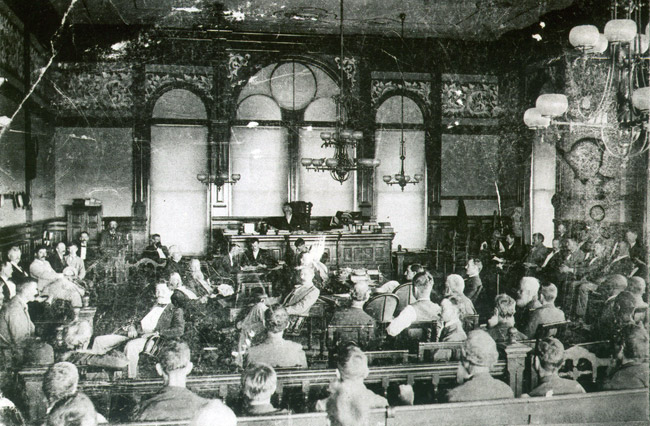 White men in crowded courtroom with arched windows and chandeliers