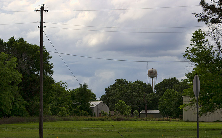 Water tower in distance with buildings on grass covered field with trees