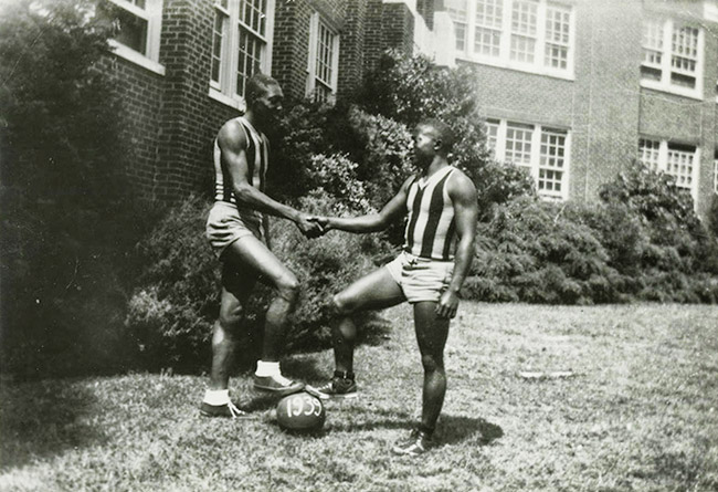 African American men in athletic wear with their feet on a ball with "nineteen thirty-nine" painted on it shaking hands outside multistory brick building