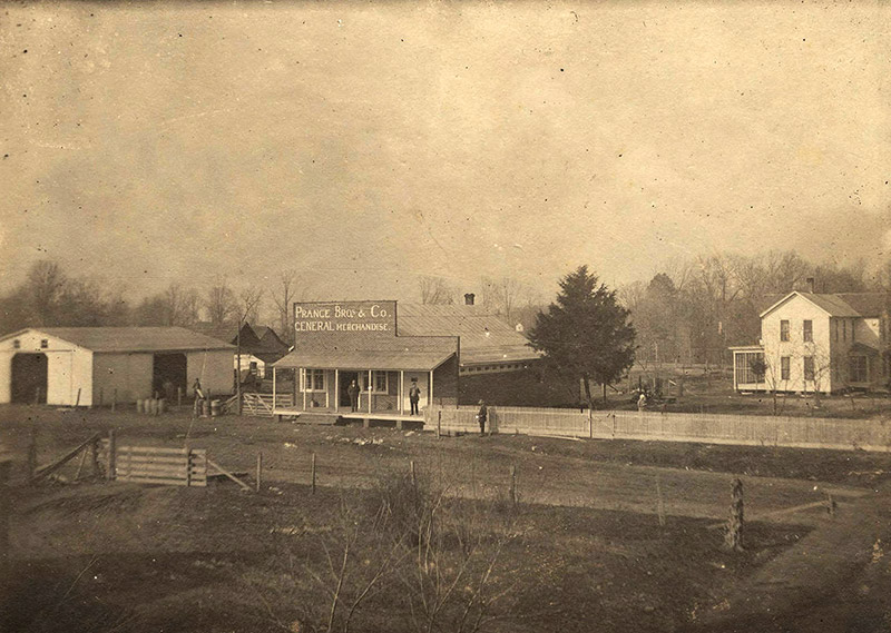 Barn and storefront with covered entrance on dirt road with fence and multistory house behind it