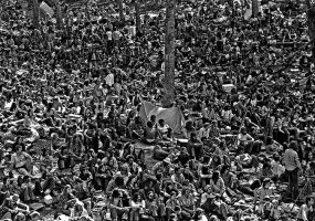 Large crowd sitting close together with tents and tree trunks