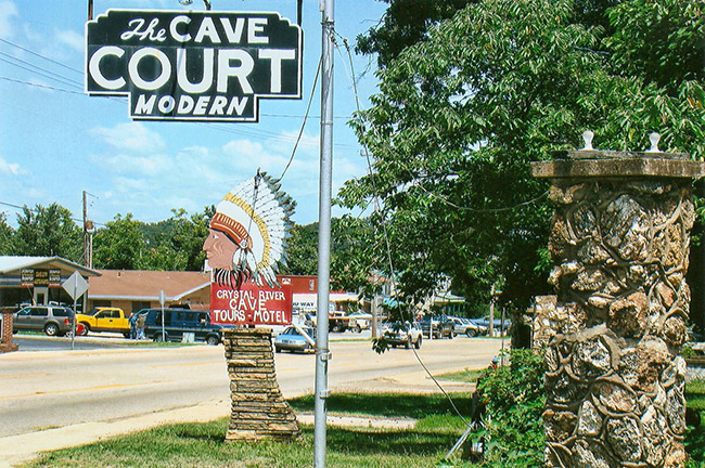 "The Cave Court modern" hanging sign and sign featuring head of Native American in feathered head dress on multilane street with storefronts in the background