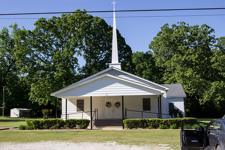 Single-story church building with white siding covered entrance and tall steeple with cross