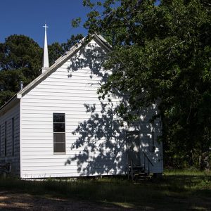 Single-story church building with white siding and steeple on dirt road