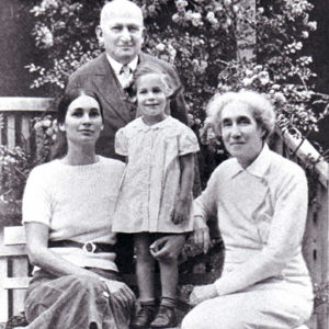 Old white man in suit standing behind bench with his wife and daughter sitting on it with little girl standing in between them