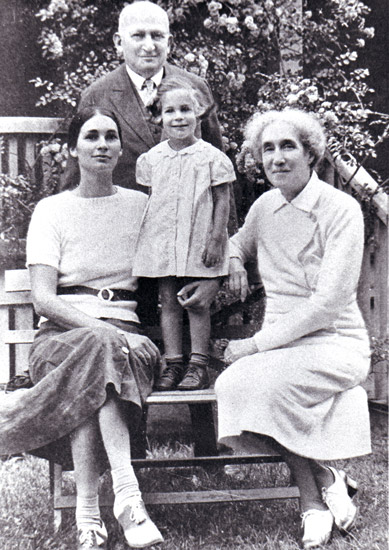 Old white man in suit standing behind bench with his wife and daughter sitting on it with little girl standing in between them
