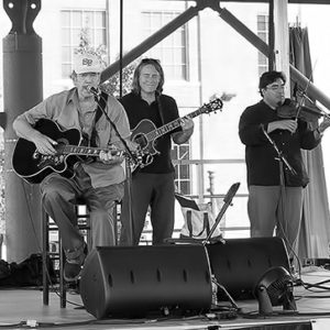 Two white woman and three white men performing music on outdoor stage with multistory building behind them