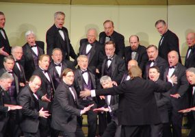 Chorus and orchestrator on stage white men in tuxedos purple vests singing with jazz hands