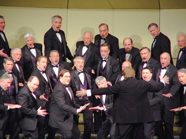 Chorus and orchestrator on stage white men in tuxedos purple vests singing with jazz hands