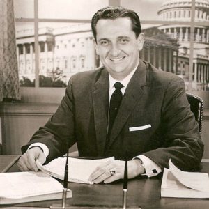 White man in suit and tie sitting at his desk with flag and U.S. Capitol visible in the background through a window