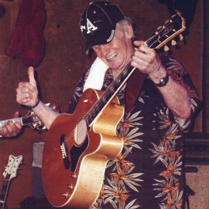 White man in ball cap and Hawaiian shirt playing acoustic guitar on stage