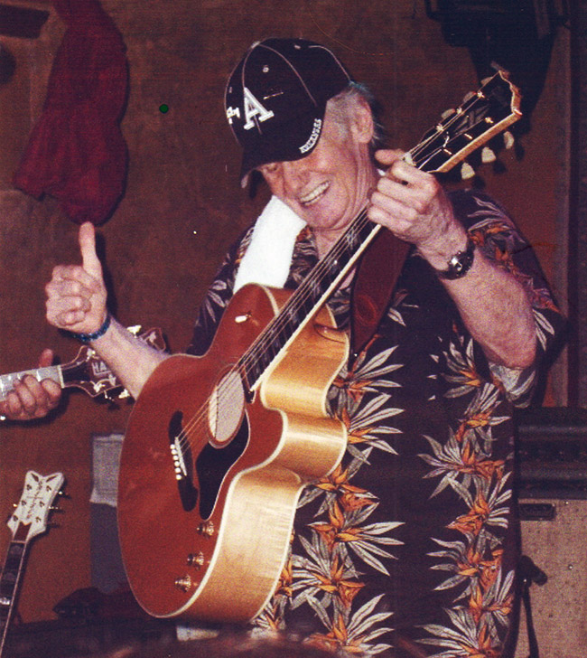 White man in ball cap and Hawaiian shirt playing acoustic guitar on stage