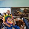 White man in tie dye shirt in wheelchair with dog in kennel and desk behind him