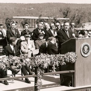 White in suit man speaking behind a lectern outdoors with crowd of officials in both military and civilian suits seated behind him
