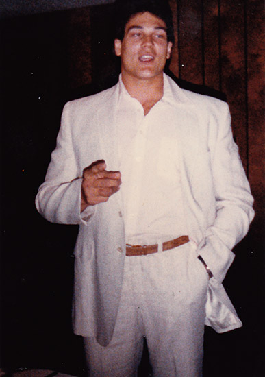 White man standing and gesturing with his right hand in white suit and brown belt