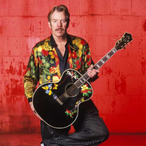 White man with mustache in colorful shirt and striped pants holding an acoustic guitar with flowers painted on it in front of red background
