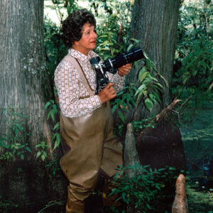 White woman with curly hair wearing waders and holding a camera with telephoto lens in a swamp