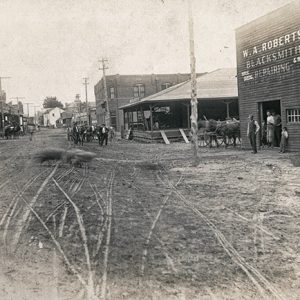 White patrons outside blacksmith shop and horse drawn wagon on dirt road with power or telephone lines and storefronts on both sides