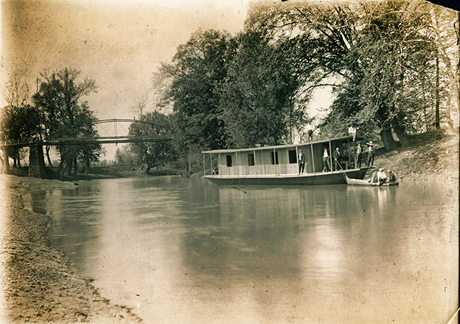 Men on houseboat on river with trees and steel arch bridge in the background