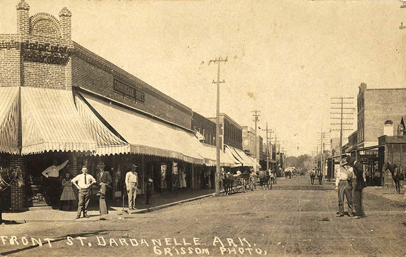 Townspeople and horse drawn wagons on town street lined with storefronts