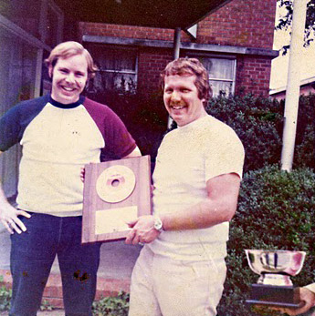 Two white men with mustaches posing with award outside brick building