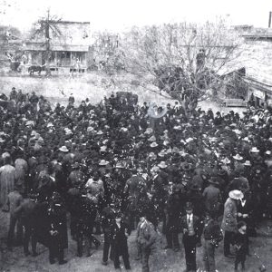 Large crowd gathered by tree with town buildings around them