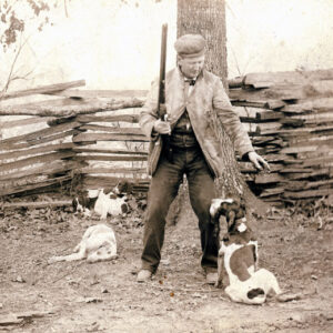 White man in outdoor clothing with gun and hunting dogs in front of wooden fence