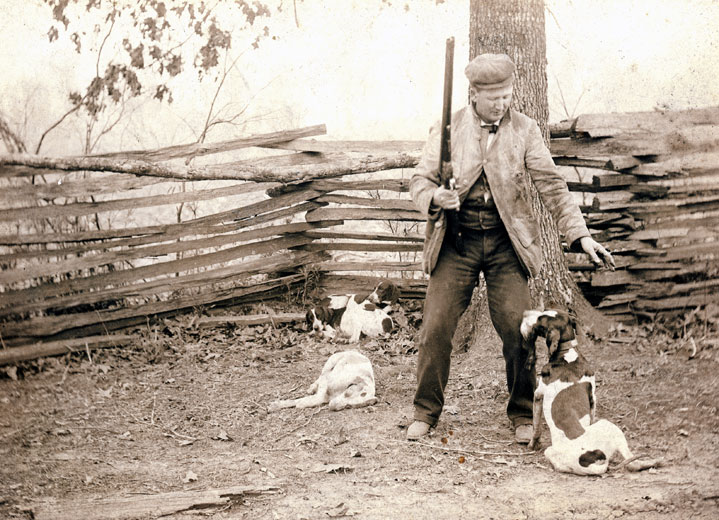 White man in outdoor clothing with gun and hunting dogs in front of wooden fence