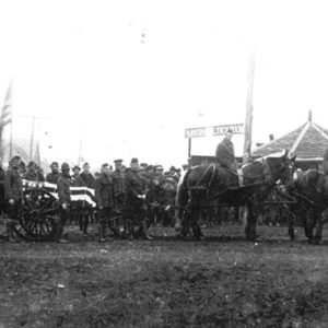 Parade of men in military uniform standing behind a horse drawn flag draped funeral wagon