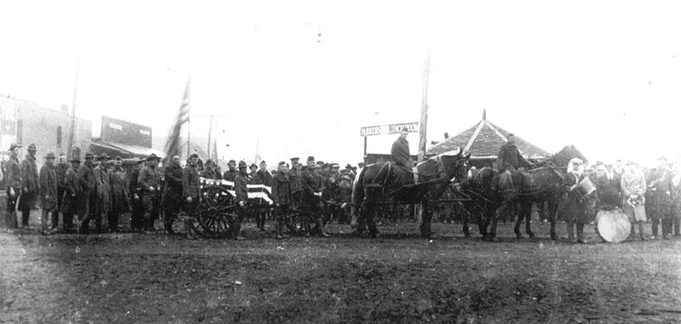 Parade of men in military uniform standing behind a horse drawn flag draped funeral wagon