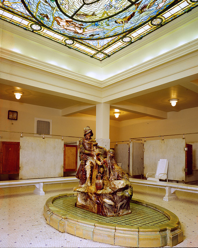 Sculpture of Native American woman pouring water for man in armor on oval base in bathing room with multiple stalls and stained glass ceiling