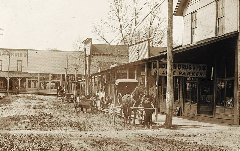Horse drawn carriage and wagon on dirt street outside storefront buildings with covered entrances