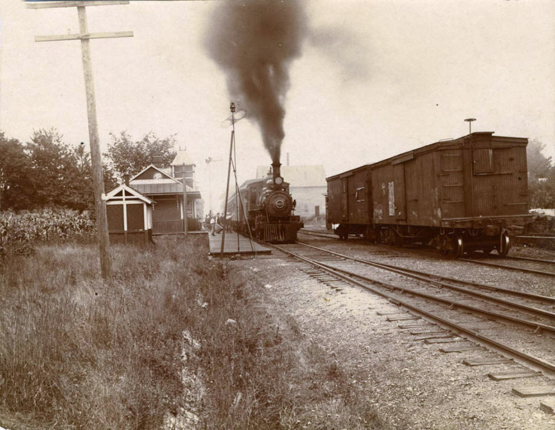 Steam train arriving at depot building with box car on separate track in the foreground