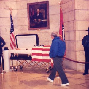 Older white man views open casket under painting with flags and police guard in State Capitol building