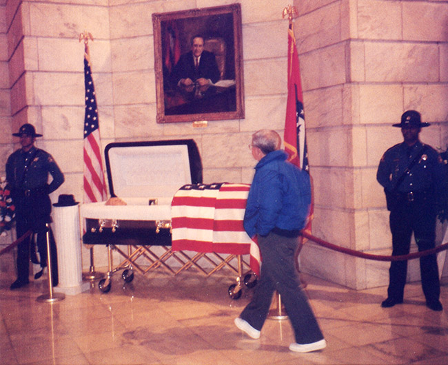 Older white man views open casket under painting with flags and police guard in State Capitol building