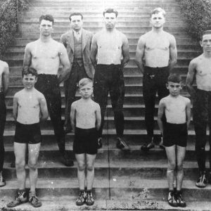 Group of young white men and boys posing shirtless on steps with white man in suit